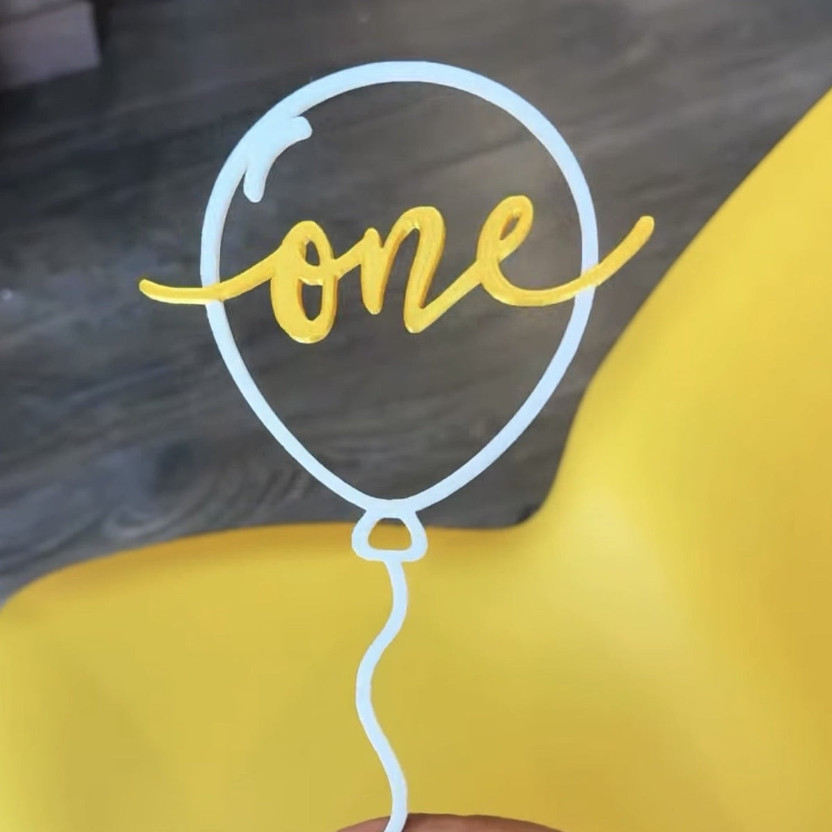 Personalized balloon cake topper — white balloon outline with gold script word 'one', held in hand